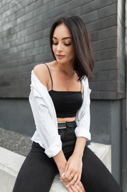 Seksuele stedelijke brunette mooie vrouw met zwarte top en spijkerbroek met wit overhemd zit op straat in de buurt van een zwarte bakstenen muur