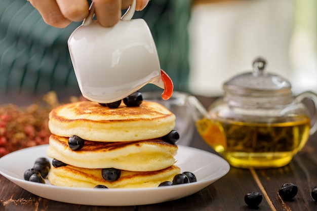 Selcetive focus op aardbeienjam gieten op pannenkoek met bosbessen fruit in witte plaat op theepot achtergrond.