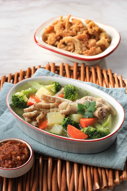 Selected focus piedi di pollo (ceker) su zuppa chiara di verdure con patate, broccoli e carote. servito su un tavolo bianco in una ciotola marrone con sambal e funghi ostrica croccanti.