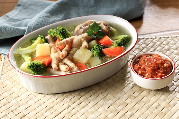 Selected focus chicken feet (ceker) on vegetable clear soup with potato, broccoli, and carrots. served on wooden table in brown bowl with sambal. copy space for text