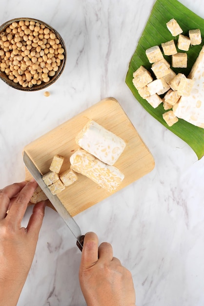 Selected focus female hand slicing cut tempeh or tempe on\
wooden chopping board using knife tempe is fermented soy product\
originally from indonesia cooking process making stir fry tempeh\
top view