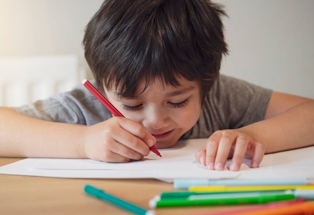 Selectieve aandacht van schooljongen jongen aanbrengen op tafel huiswerk, gelukkig kind met rode pen schrijven of tekenen op wit papier, basisschool en thuisonderwijs, onderwijsconcept