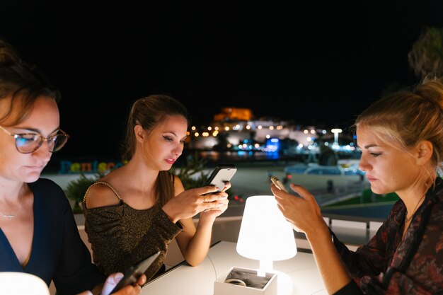 Selectieve aandacht voor een jonge vrouw zittend op een terras met andere vrouw 's nachts tijdens het gebruik van hun mobiele telefoon