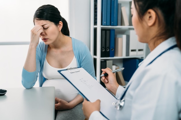 Selectieve focus depressieve zwangere moeder steunt gezicht in wanhoop op het bureau terwijl de vrouwelijke gynaecoloog op klembord schrijft tijdens consultatie in de kliniek