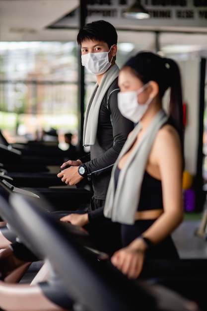 Selectieve focus jonge man met masker, wazige jonge sexy vrouw op de voorgrond die sportkleding en smartwatch draagt, ze staan een programma op de loopband in te stellen om te trainen in een moderne sportschool, kopieer ruimte