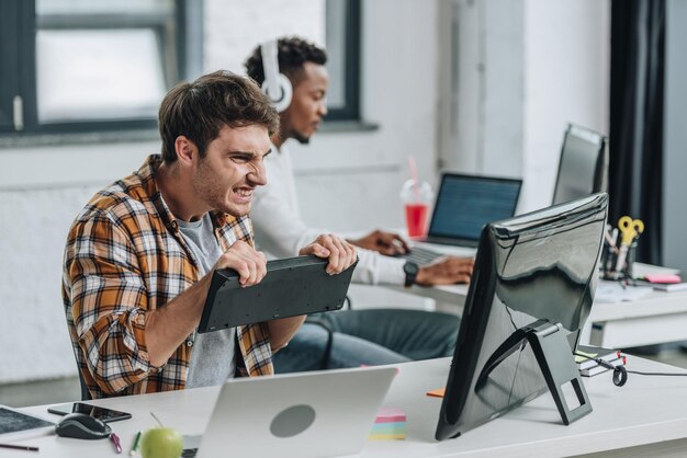 Foto selectieve focus van een boze programmeur die het toetsenbord vasthoudt en naar de computermonitor kijkt terwijl hij zit