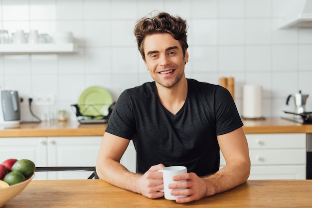 Selectieve focus van een jonge man die lacht naar de camera terwijl hij een koffiekopje vasthoudt aan tafel in de keuken