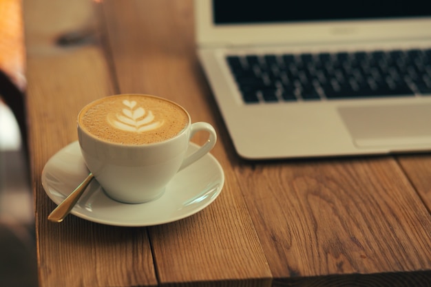 Selectieve focus van een witte kop koffie op een bord. Moderne laptop op de achtergrond achter de beker