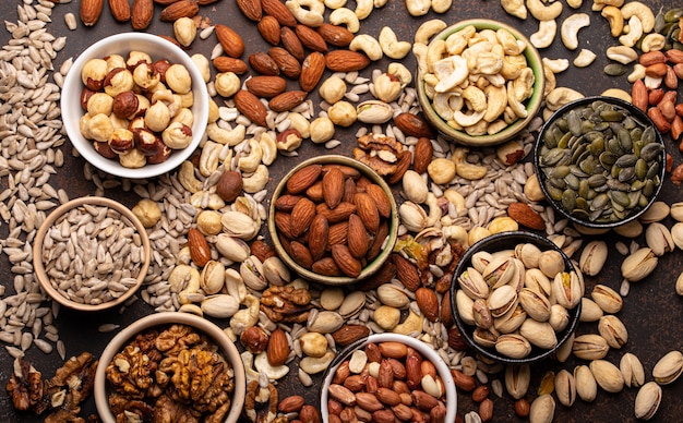 Selection of assorted raw nuts and various seeds in bowls on brown stone background from above, healthy source of energy, fat and vegetarian protein