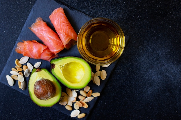 Selection of healthy fat sources, avocado, salmon, nuts, olive oil on a black plate. The concept of healthy eating. Top view, copy space, black background