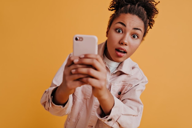 Selective focus of amazed black woman with smartphone Curious african american girl holding digital device isolated on orange background