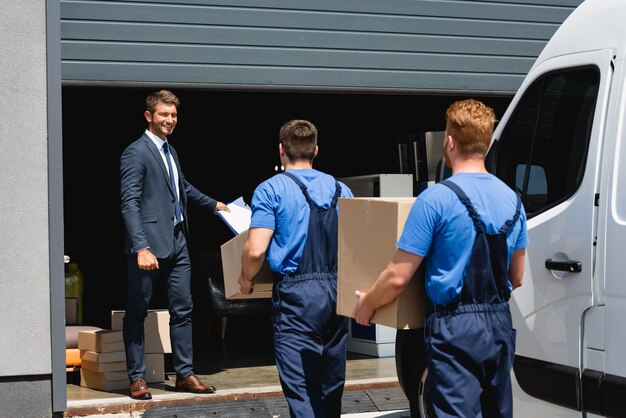 selective focus of businessman holding clipboard while movers carrying packages near truck and