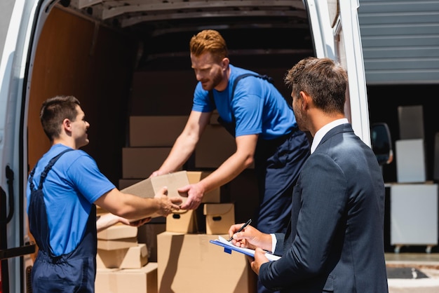 Photo selective focus of businessman holding pen and clipboard while movers unloading packages from truck