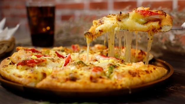 Selective focus and close up on a piece of double cheese homemade seafood pizza on spatula or scoop with blur background of wooden table, cold beverage and brick wall. Food Concept.