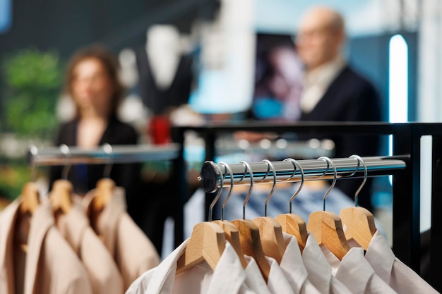 Selective focus of clothes on hangers in modern boutique, in background customers shopping for casual wear. Clients buying fashionable clothes and trendy accessories in clothing store