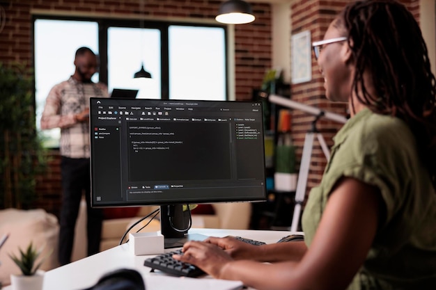 Selective focus on computer screen with programming language text of programmer working remote in home living room. African american coder developing code on pc while boyfriend is using laptop.