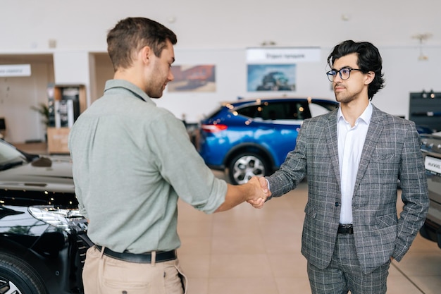 Foto focus selettivo di un concessionario di automobili sicuro di sé in abito da lavoro che accoglie un giovane cliente nello showroom