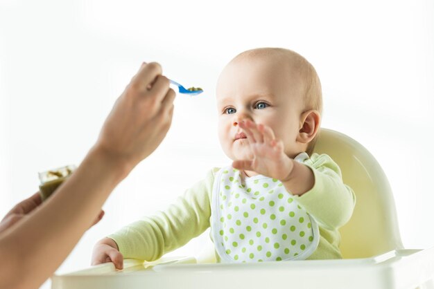 Selective focus of cute baby pulling hand to mother with spoon of baby nutrition isolated on white