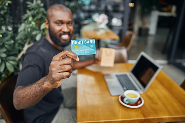 Selective focus of a debit card in hand of a smiling bearded Afro American man at the cafe table