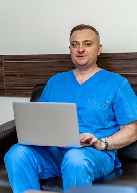 Selective focus on doctor in blue scrubs. Sitting on sofa with laptop.