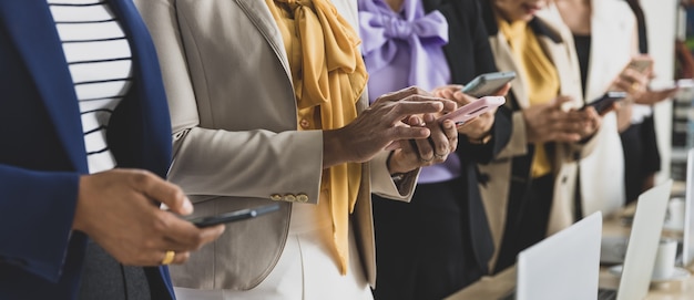 Selective focus on five Asian business women in formal suits torso standing together in office using smartphones.