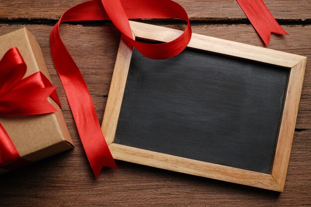 Selective focus of gift box with red ribbon, chalkboard and love shaped paper over rustic wooden table, top view flat lay.