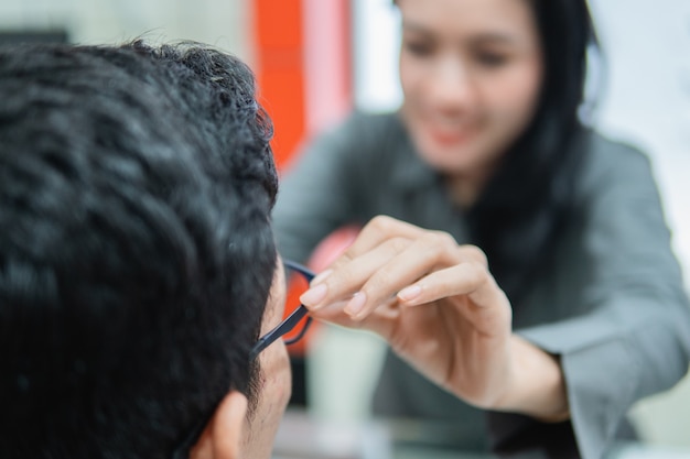 Selective focus of the hand of woman shop assistant helps a man put on glasses while sitting trying on glasses in an optician