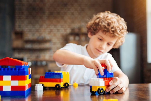 Selective focus on hands of a smart curly haired boy sitting at a table and designing his dream car while playing with a construction set at home.