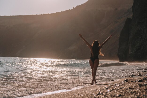 サンセットビーチでポーズをとる黒い水着で長い髪の選択的な焦点幸せなのんき官能的な女性
