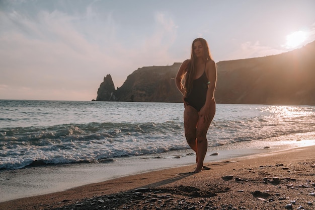 Donna sensuale spensierata felice del fuoco selettivo con i capelli lunghi in costume da bagno nero che posa alla spiaggia del tramonto