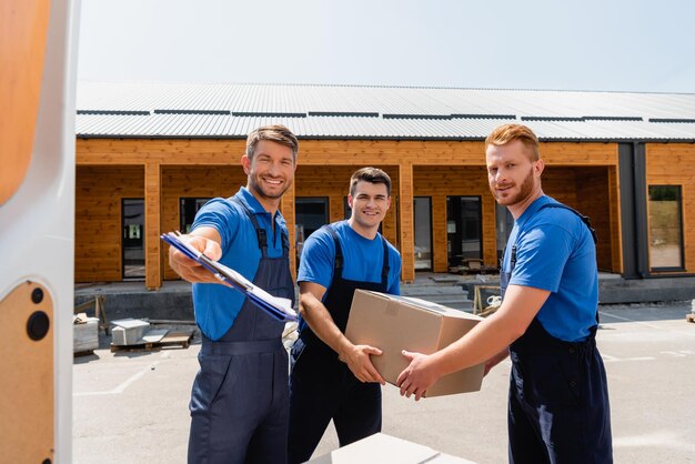 Selective focus of loader holding clipboard near colleagues with cardboard box and door of truck on