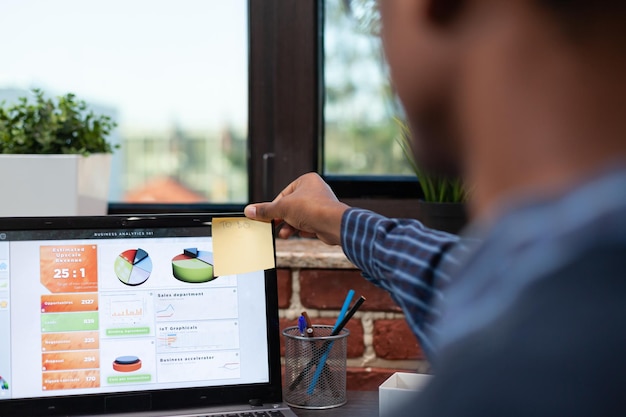 Selective focus on marketing specialist hand sticking to do note in corner of laptop screen with business analytics data. Closeup view of business analyst putting reminder post it on portable computer