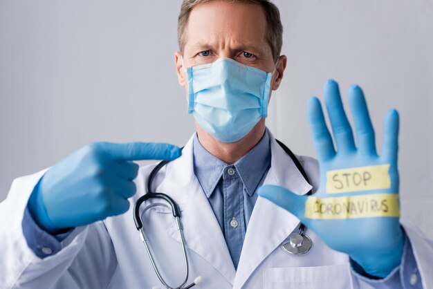 Selective focus of mature doctor in medical mask pointing with finger at hand with stop coronavirus