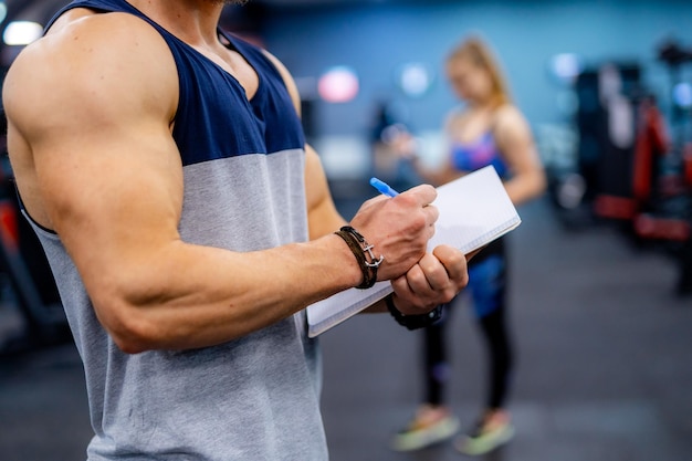 Photo selective focus on muscular hands with clipboard. unrecognizible fitness trainer gives recommendations for better results. client girl on blurred background.