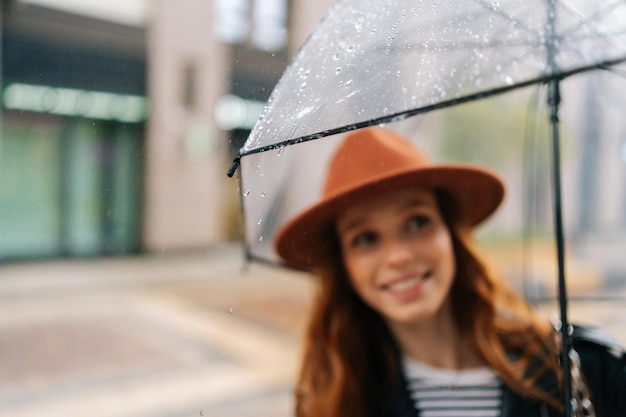 写真 屋外で雨天を楽しんでいる街の通りに透明な傘を持って立っている帽子をかぶった幸せな若い女性の選択的な焦点