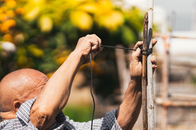 Selective focus on a old man tying a rope in a wooden stick structure in a orchard