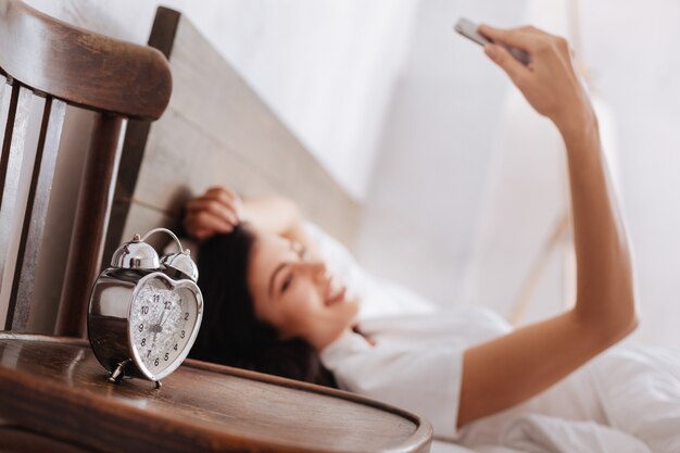 写真 ベッドに横たわっている間に自画像を撮る女性と一緒に木製の椅子に立っている銀の目覚まし時計に選択的な焦点