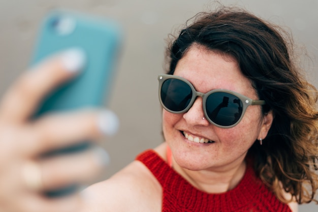 Selective focus of a portrait of smiling woman wearing sunglasses and using her phone to talk on a video call.