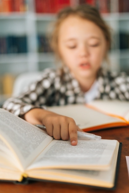 テーブルに座って紙の本を読んで宿題をしているかわいい生徒の女子高生の子供の選択的な焦点