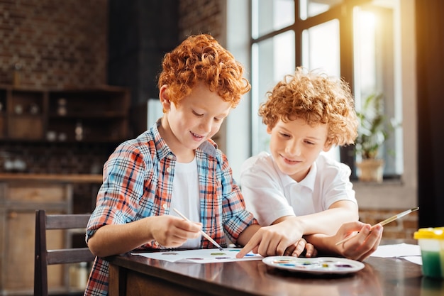 Selective focus on a redhead child wearing a plaid shirt listening to his older brother giving him advice and pointing at a piece of paper while both painting at home.