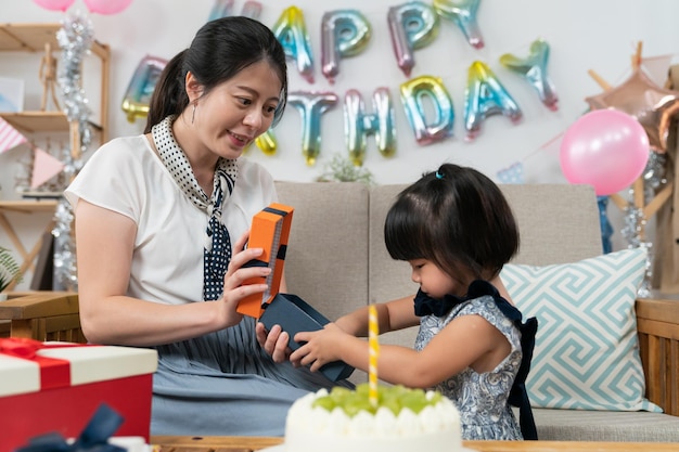 selective focus of smiling asian mother opening and showing her baby daughter present as the curious girl's hand reaching inside the box at birthday party at home