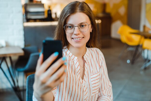 Selective focus of smiling girl in glasses taking selfie on smartphone in cafe