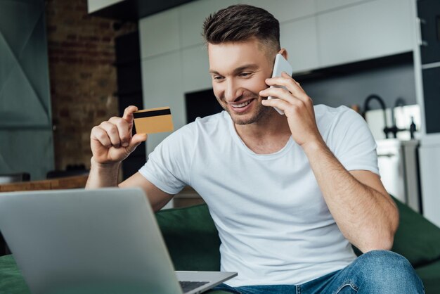 Selective focus of smiling man holding credit card while talking on smartphone near laptop in living