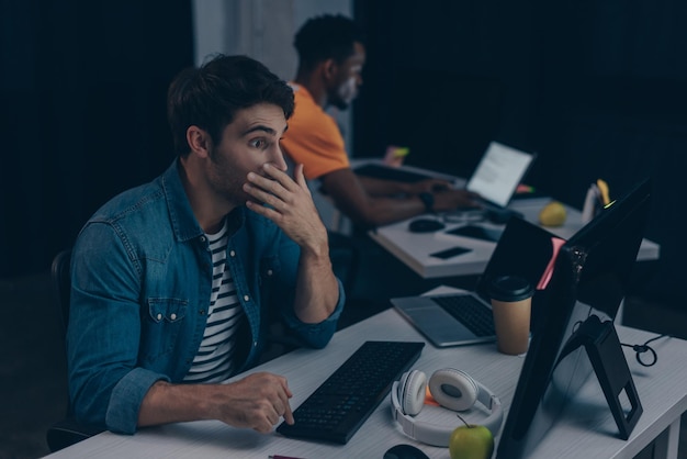 Selective focus of surprised programmer looking at monitor while working near african american