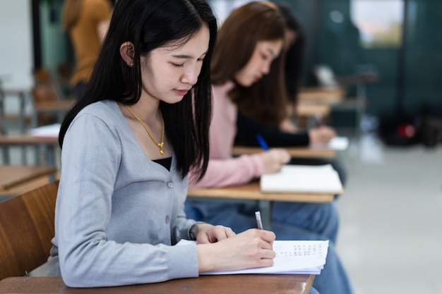 Selective focus of the teen college students sitting on lecture\
chair in classroom write on examination paper answer sheet in doing\
the final examination test. female students in the student\
uniform.