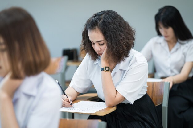 Selective focus of teenage college students sit on lecture chaire, write on examination paper answer sheet taking final examination room or classroom. University students in uniform in classroom.