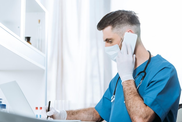 Selective focus of young doctor in medical mask and latex gloves writing while talking on smartphone