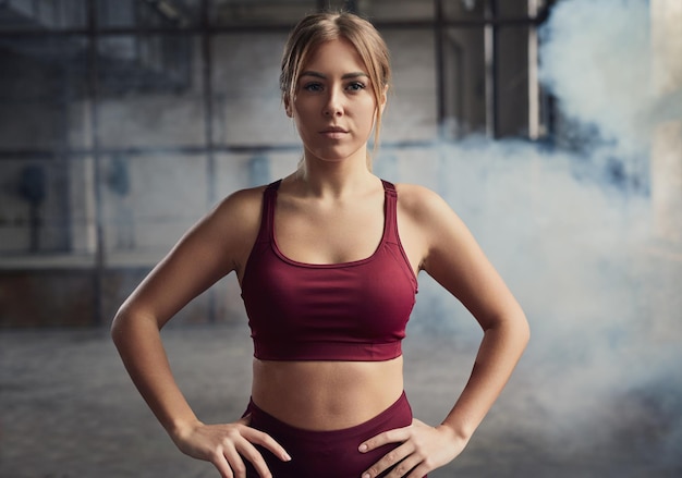 Self assured young fit female athlete in sportswear with hands on hips looking forward against dust in gymnasium