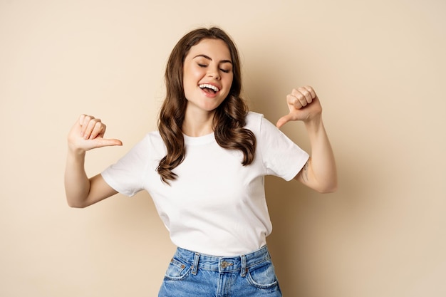 Self-assured young happy woman pointing fingers at herself and dancing, self-promoting, being confident, standing over beige background