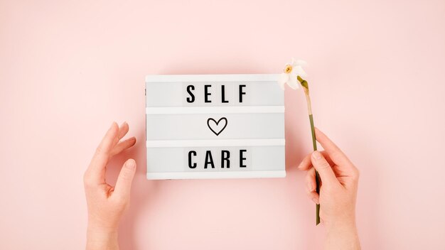 Photo self-care word on lightbox and flower narcissus on pink background flat lay take care of